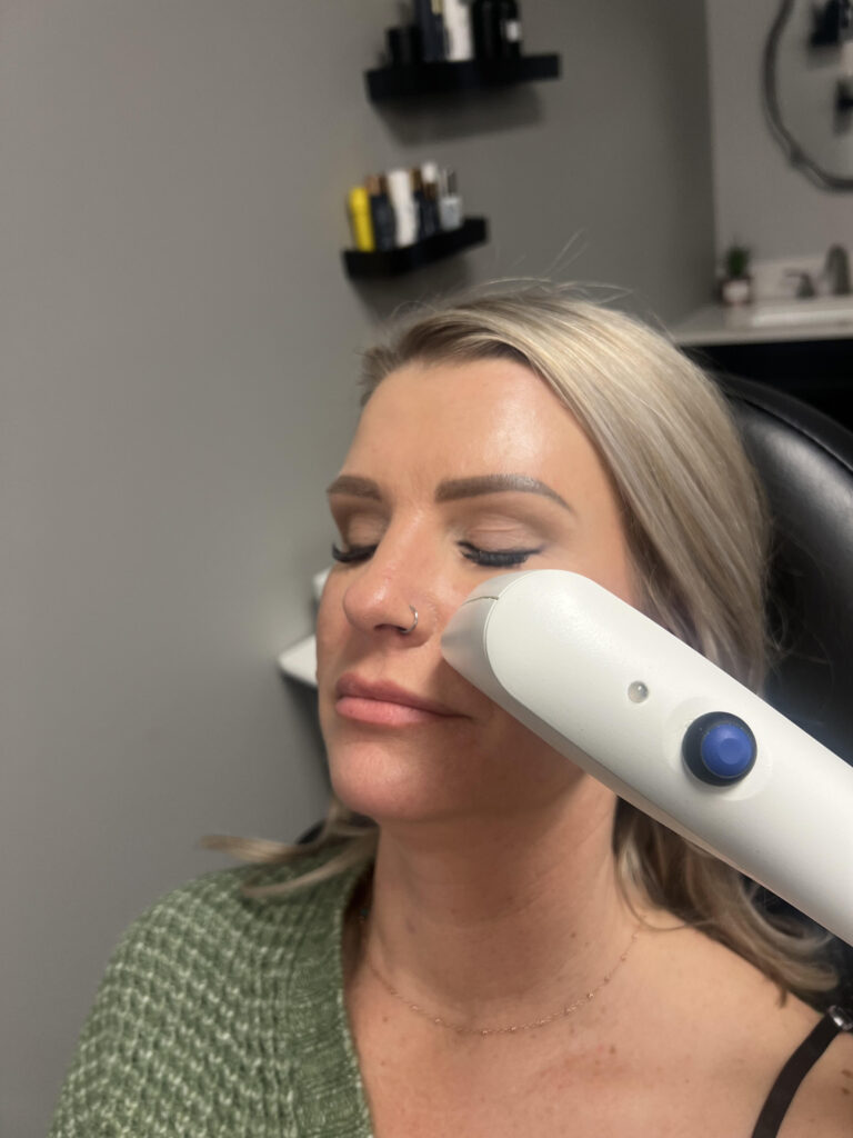 A woman with closed eyes undergoes a skin treatment using a handheld device on her face. She appears relaxed, wearing a green sweater. Shelves with products are in the background.